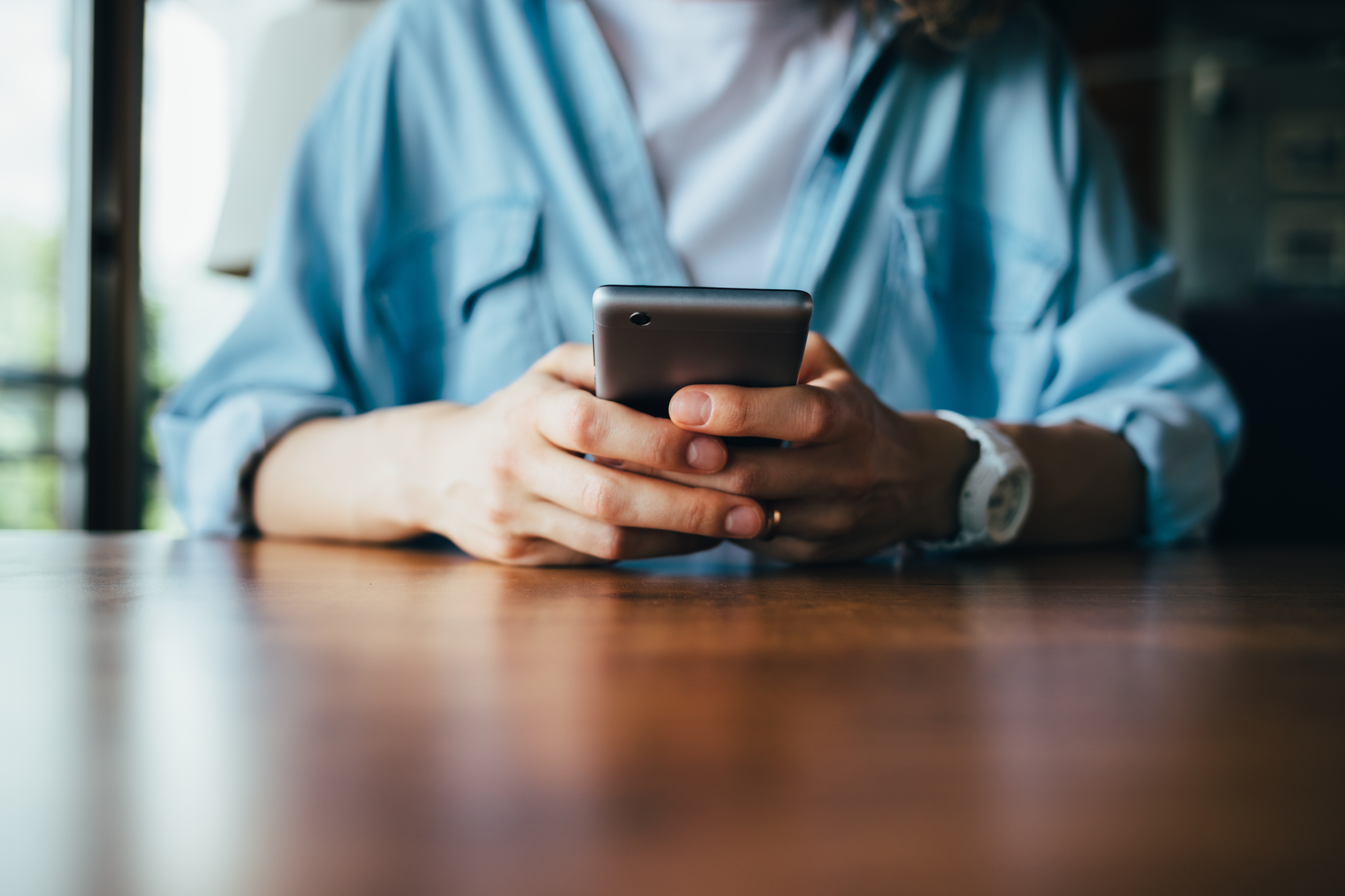 Close-up of a person holding a smartphone with both hands, sitting at a wooden table. The person is wearing a light blue button-up shirt over a white t-shirt and a white wristwatch.